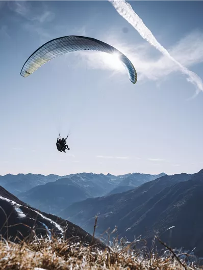 A paraglider is floating over a mountain landscape. The sky is clear and the mountains are visible in the background.