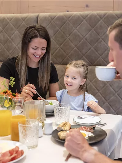 Een gezin geniet van een ontbijt aan de tafel. Het meisje glimlacht gelukkig, terwijl de volwassenen met eten bezig zijn.