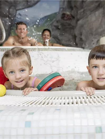 Two cheerful children are playing in the water of a kiddie pool. In the background, there are adults who are also having fun.