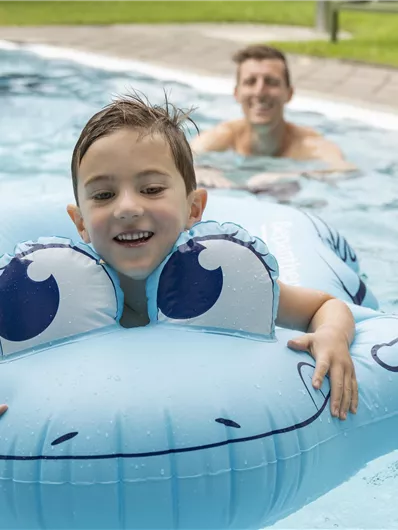 A cheerful boy with a blue frog float swims in the pool. In the background, a smiling man can be seen.