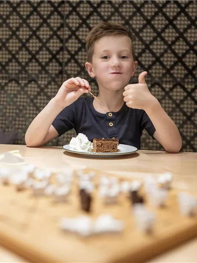 A boy is sitting at a table and enjoying a piece of cake with cream. He is giving a thumbs-up sign.