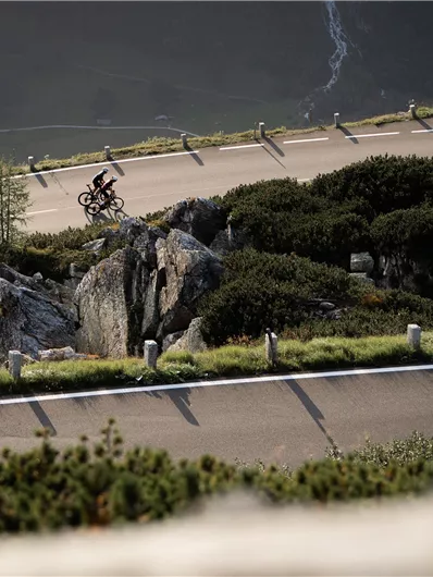 A cyclist rides along a winding road in a mountainous landscape. Surrounded by rocks and green plants, the scene conveys a sense of adventure.
