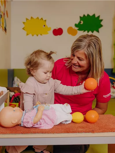 Ein kleines Kind spielt mit einer Frau in einem bunten Spielzimmer. Auf dem Tisch liegen Spielzeugfrüchte und eine Puppe.