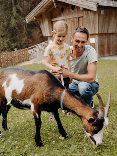 Ein Vater spielt mit seiner Tochter auf einer Wiese. Eine Ziege grast in der Nähe eines Holzhauses.