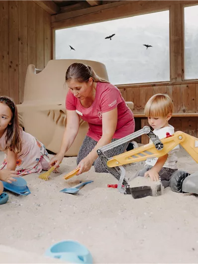 Eine Gruppe von Kindern spielt im Sandkasten mit Spielsachen, während eine Aufsichtsperson ihnen hilft. Es gibt einen großen Geländewagen und bunte Schaufeln.