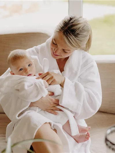 Eine Mutter sitzt auf einem Sofa und hält ihr Baby in den Armen. Beide tragen weiße Bademäntel und wirken entspannt in einem hellen Raum.