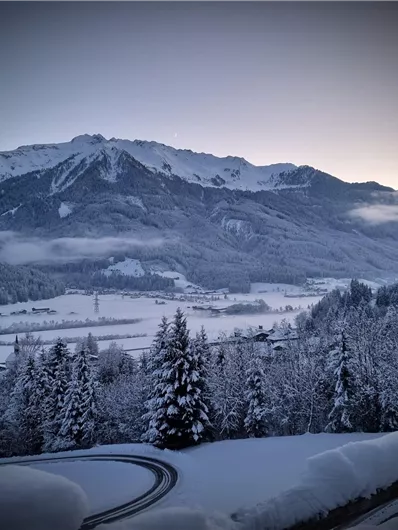 A snowy mountainous landscape with tall mountains and snow-covered trees. The sky is clear and displays the colors of the sunrise.
