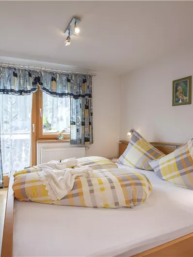 A cozy bedroom with a double bed and light wooden furniture. The curtains and windows let in plenty of light.