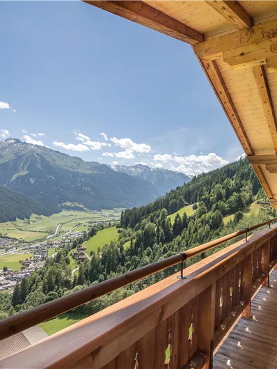 A balcony with wooden cladding and two chairs that offers a stunning view of the surrounding mountains and the green valley. The sky is clear with some clouds.