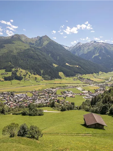 A picturesque Alpine landscape with green meadows and mountains in the background. In the foreground, there is a small wooden house and a view of the village in the valley.