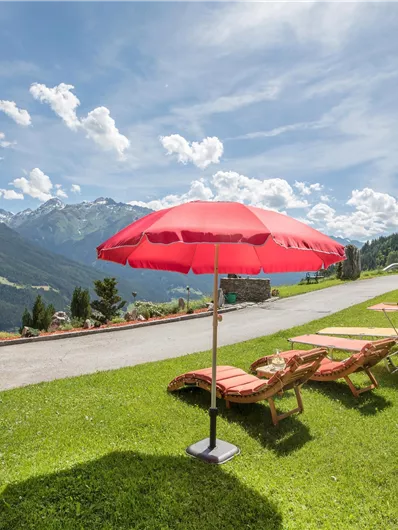 A beautiful, sunny garden with deck chairs and a red parasol. In the background, gentle hills and a picturesque mountain landscape can be seen.
