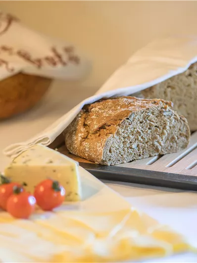 A freshly baked bread, partially peeking out from the cloth. Next to it are various types of cheese and some cherry tomatoes on a plate.