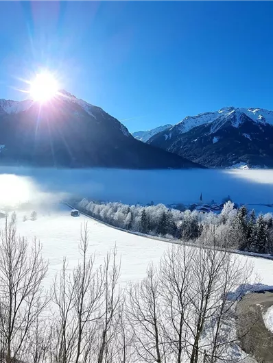 A winter landscape with snow-covered fields and mountains in the background. The sun shines clearly over the scene, creating a picturesque atmosphere.