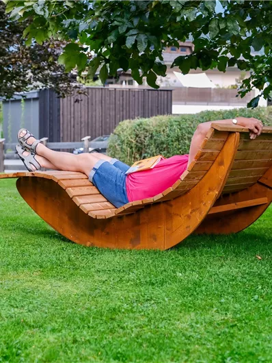 A person is relaxing on a modern wooden lounger in a green area. In the background, there are trees and a well-maintained lawn.