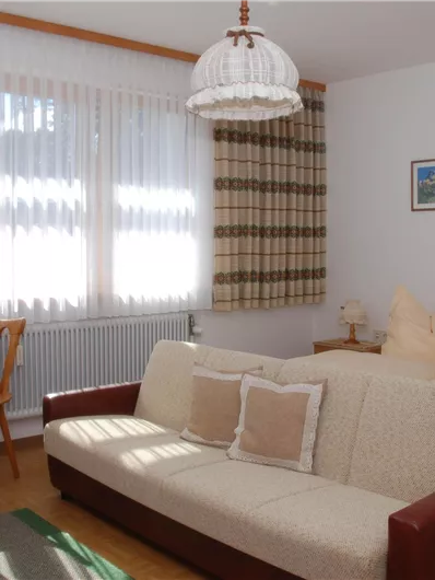 Ein gemütliches Schlafzimmer mit zwei Betten, einem Sofa und einem Tisch. Große Fenster lassen viel Licht hinein und schaffen eine einladende Atmosphäre.