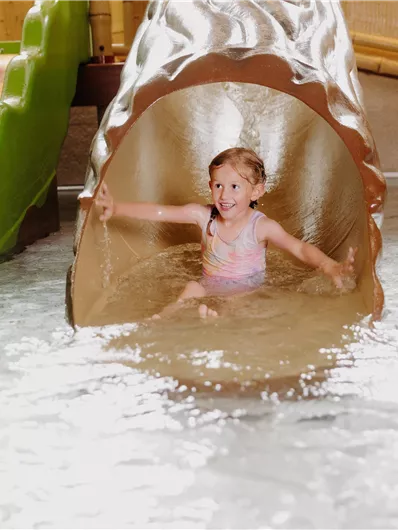 Ein fröhliches Kind rutscht von einer Wasserbahn ins Wasser. Umgeben von spritzendem Wasser und bunten Rutschen hat es viel Spaß.