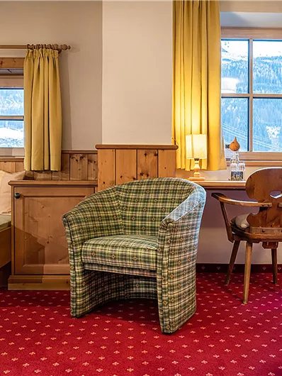 Ein gemütliches Zimmer mit Holzverkleidung, einem Sessel und einem kleinen Tisch. Die großen Fenster lassen viel Licht herein und bieten einen Blick nach draußen.