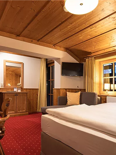 Ein gemütliches Hotelzimmer mit Holzelementen und roten Teppich. Große Fenster bieten einen Blick auf die Berge.