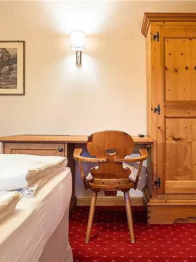 Ein gemütliches Zimmer mit einem Bett, einem Schreibtisch und einem Holzschrank. Die Wände sind in warmen Farben gehalten und der Teppich ist rot.