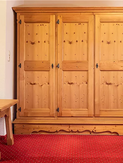 Ein klassischer Holzschrank mit drei Türen steht in einem Raum. Daneben befindet sich ein kleiner Tisch auf einem roten Teppich.