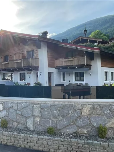 A modern house with wooden cladding and balconies, surrounded by greenery. In the foreground, a paved driveway and a stone wall can be seen.