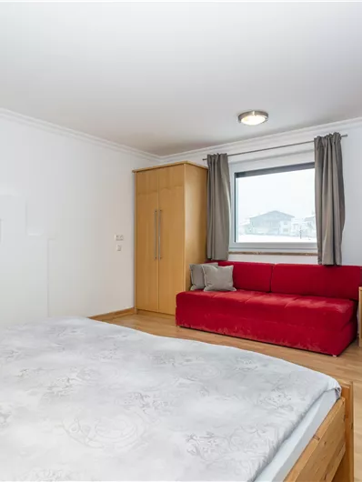 Een moderne slaapkamer met een tweepersoonsbed en een groot raam. In de kamer staan een televisie en een gezellige rode bank.