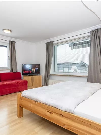 Een moderne slaapkamer met een tweepersoonsbed en grote ramen. Er is een televisie en een rode bank in de kamer.