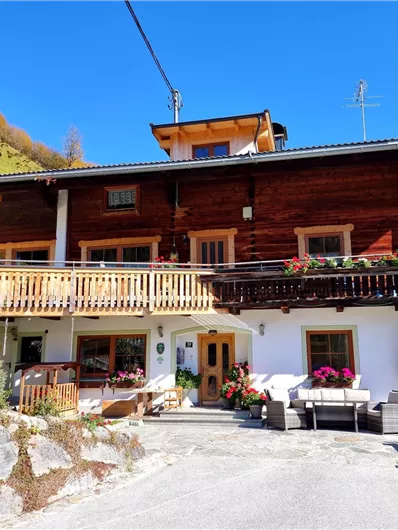 A traditional wooden house in the mountains with a large balcony and flowers. In the background, green hills and a blue sky are visible.