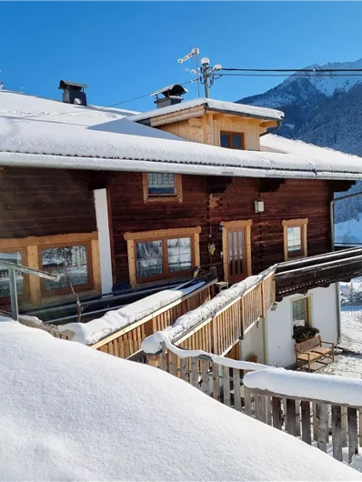 A cozy wooden house in the snow with a clear blue sky. Snow-covered mountains are visible in the background.