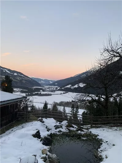 Eine winterliche Landschaft mit schneebedeckten Feldern und mehreren Bergen im Hintergrund. Der Himmel zeigt sanfte Pastellfarben bei Sonnenuntergang.