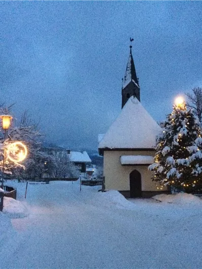 Eine verschneite Winterlandschaft mit einem kleinen Kirchturm und einem beleuchteten Weihnachtsbaum. Alltagsleben ist ruhig und friedlich in der Dämmerung.