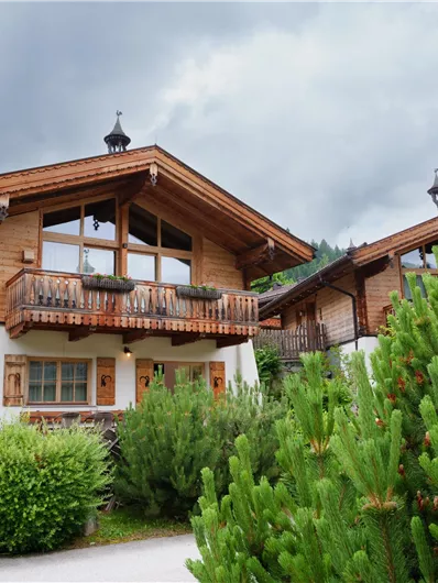 Ein gemütliches Holzhaus mit einem Balkon und großen Fenstern. Die Umgebung ist grün und von Bäumen umgeben, mit dramatischem Himmel.