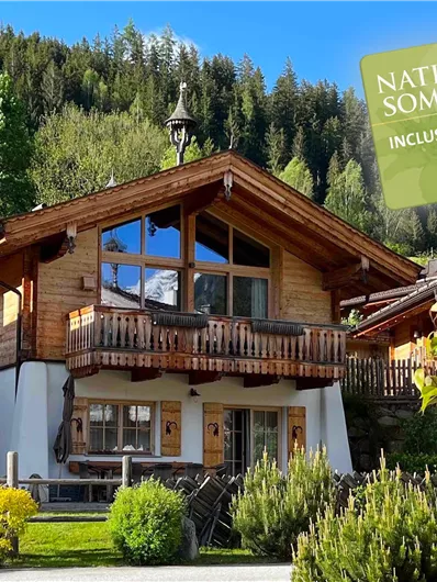 Ein charmantes Holzhaus mit Balkon, umgeben von grüner Natur und Bergen. Im Hintergrund befindet sich ein Schild mit dem Hinweis auf den Nationalpark Sommerwald.