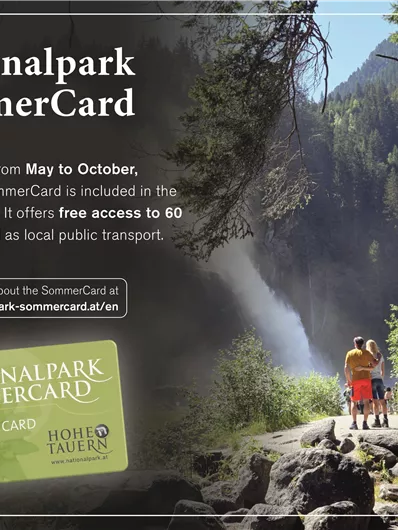 Ein Paar steht an einem Wasserfall im Nationalpark, umgeben von grünen Bäumen und einer malerischen Landschaft. Die Nationalpark SommerCard ermöglicht kostenlosen Zugang zu vielen Attraktionen und öffentlichen Verkehrsmitteln.