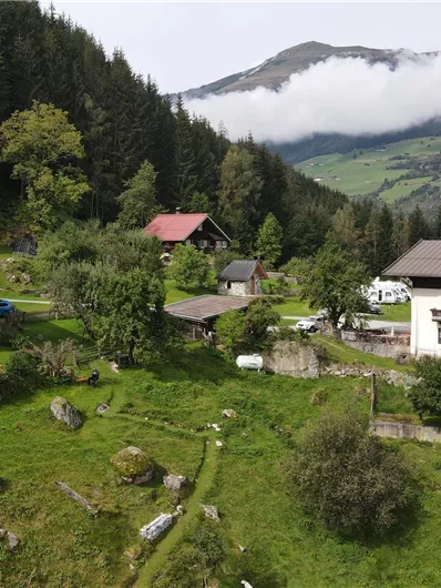 Eine malerische Almlandschaft mit mehreren traditionellen Hütten und üppigem grünen Gras. Im Hintergrund erheben sich majestätische Berge unter einem bewölkten Himmel.