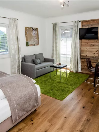 Ein modernes Wohnstudio mit einem Doppelbett, einer gemütlichen Couch und einem kleinen Essbereich. Große Fenster sorgen für viel Tageslicht und eine einladende Atmosphäre.