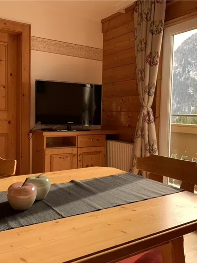 Ein gemütlicher Wohnraum mit einem Holztisch und einer TV-Einheit. Große Fenster bieten einen Blick auf die umliegenden Berge.