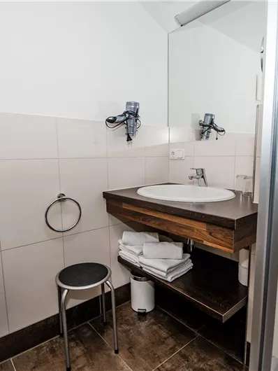 Een moderne badkamer met een wastafel, een spiegel en een douche. Er zijn handdoeken op een plank en een zitgelegenheid aanwezig.