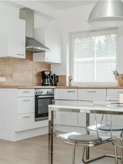 Eine moderne Küche mit weißen Möbeln und einem Essbereich aus Glas. Helle Wände und große Fenster sorgen für ein offenes Raumgefühl.