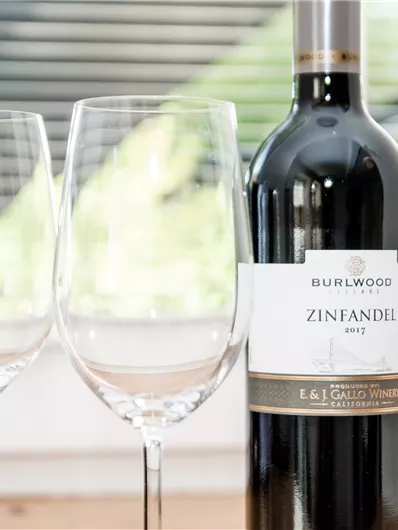 Een fles Zinfandel-wijn staat naast twee wijnglazen. De achtergrond toont ramen met zonlicht.