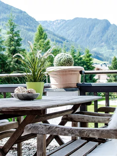 Ein gemütlicher Balkon mit einem Holztisch und Stühlen, dekoriert mit Kissen. Im Hintergrund erheben sich majestätische Berge und üppige Bäume.