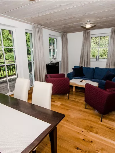 Ein helles Wohnzimmer mit einem blauen Sofa und zwei roten Sesseln. Große Fenster bieten einen Blick auf die Bäume draußen.