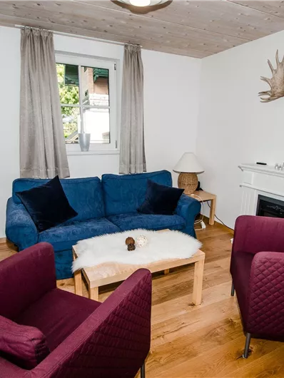 Ein gemütliches Wohnzimmer mit einem blauen Sofa und zwei roten Sesseln. An der Wand hängen Geweih-Elemente, und der Raum hat holzige Akzente.
