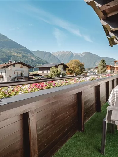A balcony with a chair and a view of a picturesque mountain landscape. The surrounding houses and flowers give the scene an inviting atmosphere.