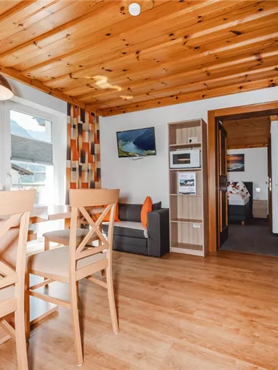 A bright and cozy holiday apartment with wooden ceilings and modern interior. The space includes a dining area, a kitchen, and access to a bedroom.