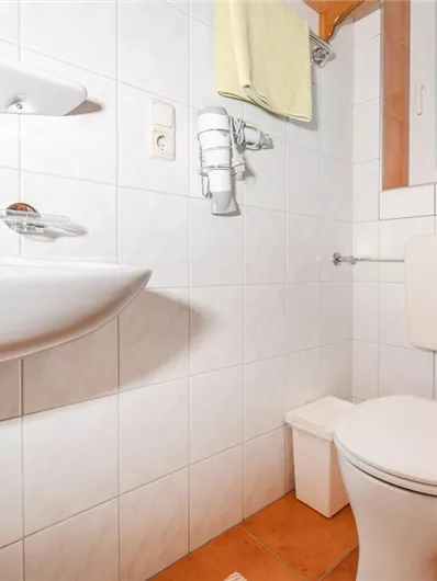 A small bathroom with a sink, a toilet, and a shower. The walls are tiled and there is a window for natural light.