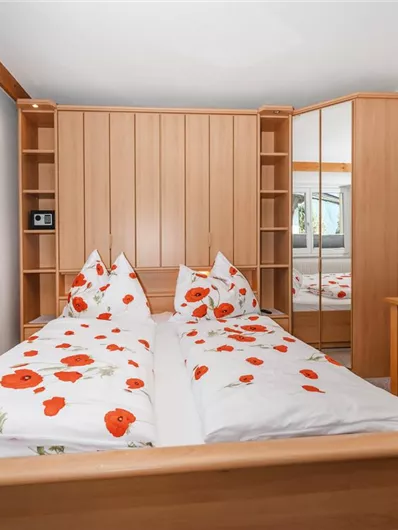 A cozy bedroom with a double bed covered in red poppy flowers. There is a table and chairs as well as a wardrobe with a mirror.