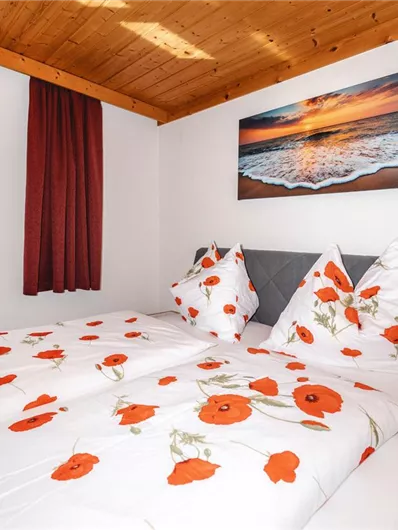 A cozy bedroom with a double bed and bedding featuring red poppies. A picture of a coastal landscape hangs on the wall, and daylight can be seen through the window.
