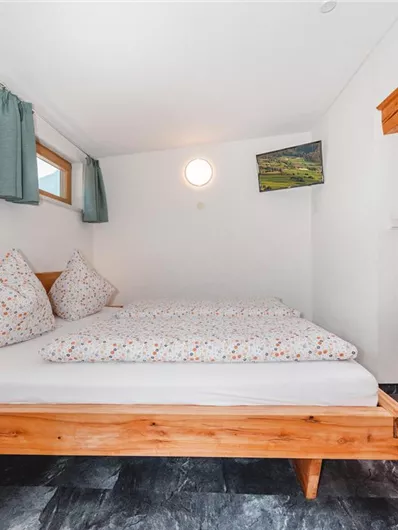 A bright bedroom with a double bed and colorful pillows. The walls are white, and there are large windows with curtains as well as a shelf.