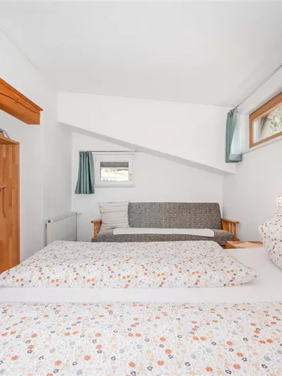 A cozy bedroom with two beds and colorful bedcovers. In the background, there is a couch and a wooden shelf.